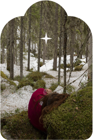 Punapaitainen pitkähiuksinen henkilö makaa lumisessa metsässä. Hän nojaa sammaleen peittämään kiveen.