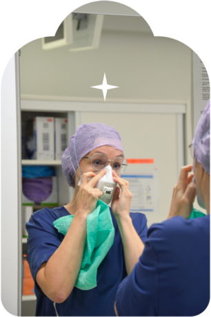 Sjuksköterskan tittar i spegeln och sätter en mask i ansiktet, ett hårnät på huvudet och en grön skyddsdräkt i handen.